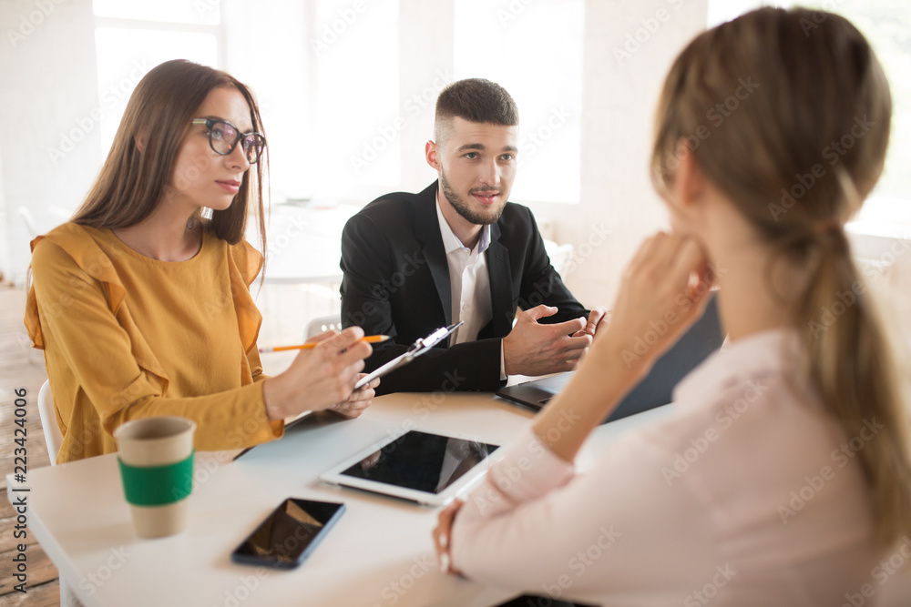 Young business man with laptop and business woman in eyeglasses with folder in hand dreamily talking with applicant about work. Thoughtful employers spending job interview in office Young business man with laptop and business woman in eyeglasses with folder in hand dreamily talking with applicant about work. Thoughtful employers spending job interview in office