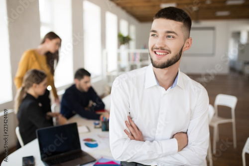 Young smiling man with beard in white shirt happily looking aside with folded hands while spending time in office with colleagues on background © Anton
