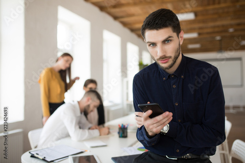 Young thoughtful business man with beard in dark shirt dremily looking in camera while using cellphone in office with colleagues on background