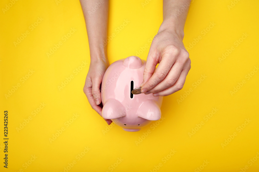Female hand putting coin into piggy bank on yellow background