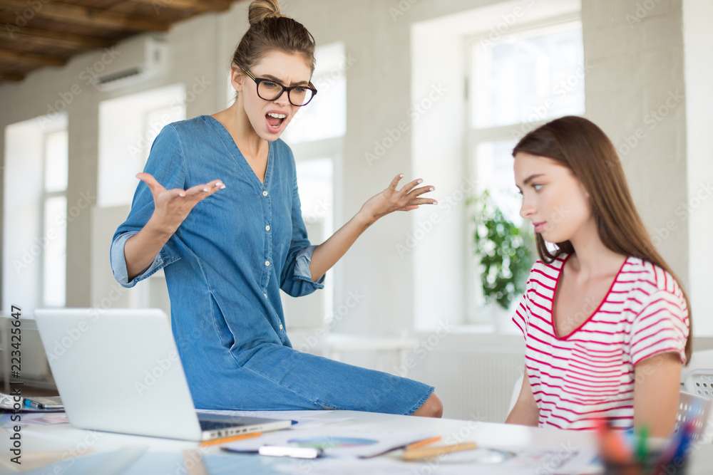 Fototapeta premium Angry business woman in eyeglasses leaning on desk while screaming on employee in modern office