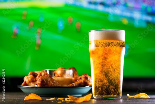 A glass of beer and hot chicken wings on a dark wooden table with crushed potato chips. Football on a background, high resolution