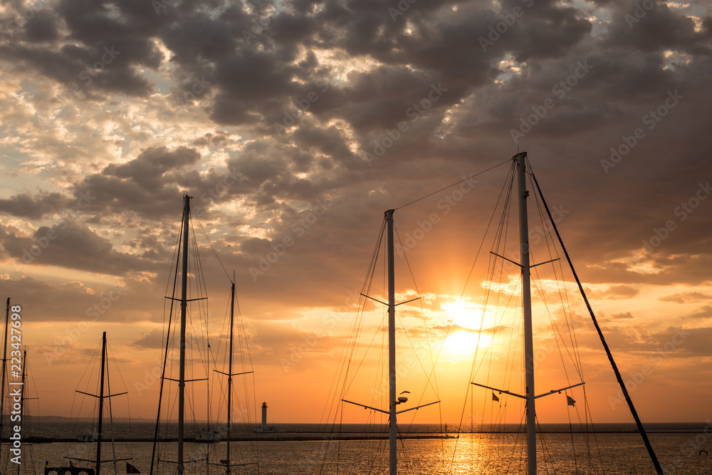 Masts of yachts in the port at sunrise against the background of the sea and the cloudy sky