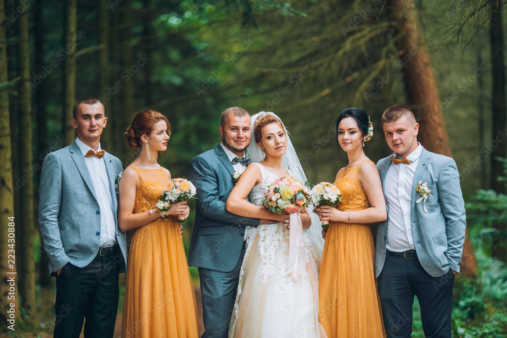 Newly married couple with groomsmen and bridesmaids posing on wedding ceremony. Bride and groom with best friends posing in pine forest. Young people having fun. Wedding day.