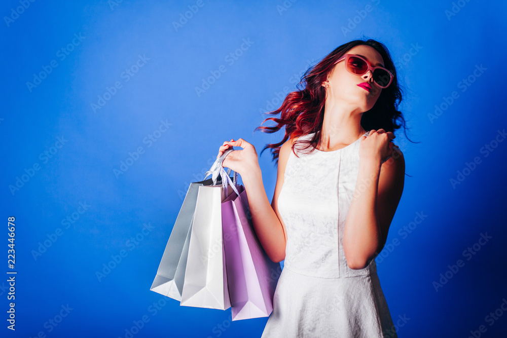 Young redhead girl in white dress with shopping bags on blue background.
