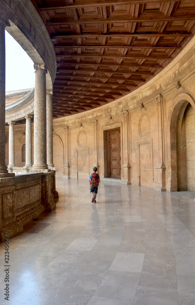 Boy traveler with backpack walking through palace hallway with wood ceiling, columns, marble floors.