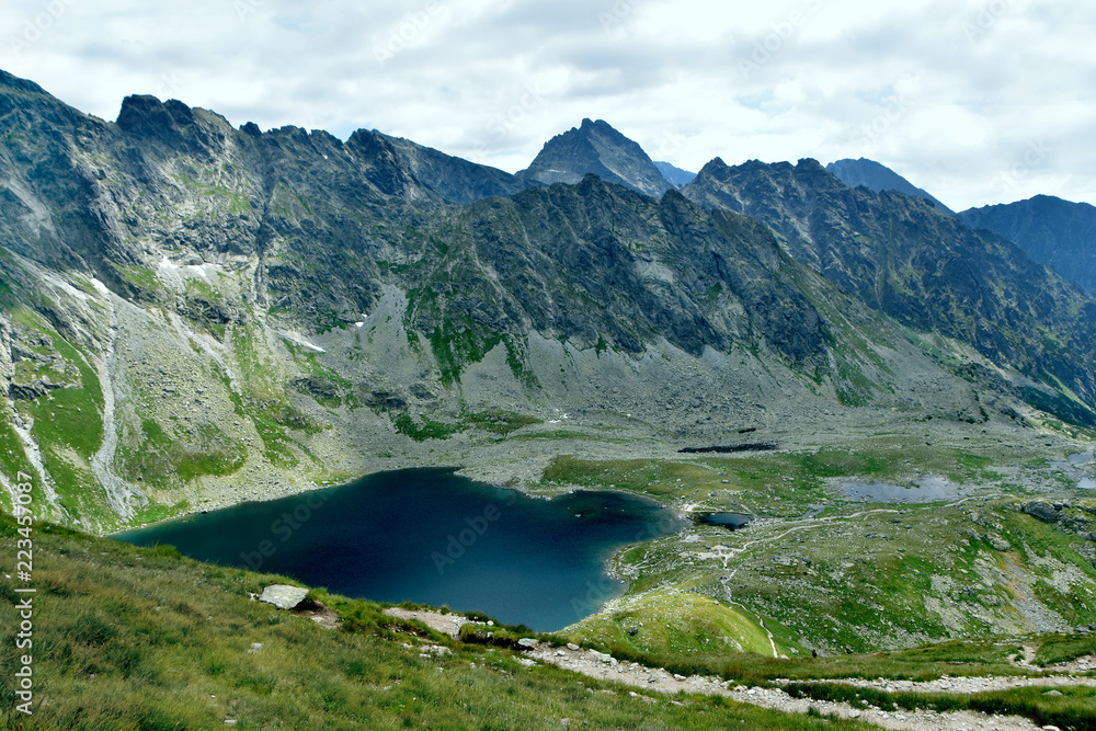 Jagged rocky peaks surrounding a deep blue glacial lake with green meadows and a gravel hiking path