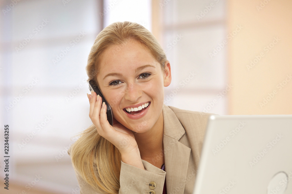 Portrait of young business girl using cell phone at desk with laptop inside office building
