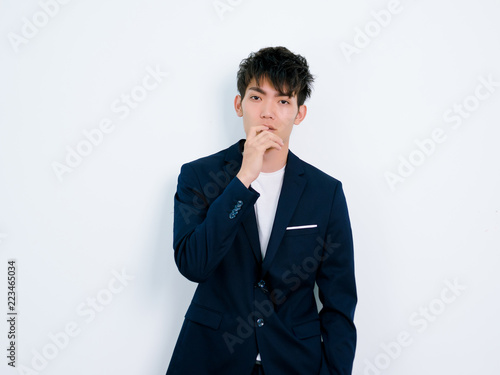 Portrait of handsome Chinese young man in dark blue leisure suit posing against white wall background. Hand on lips and looks like thinking.