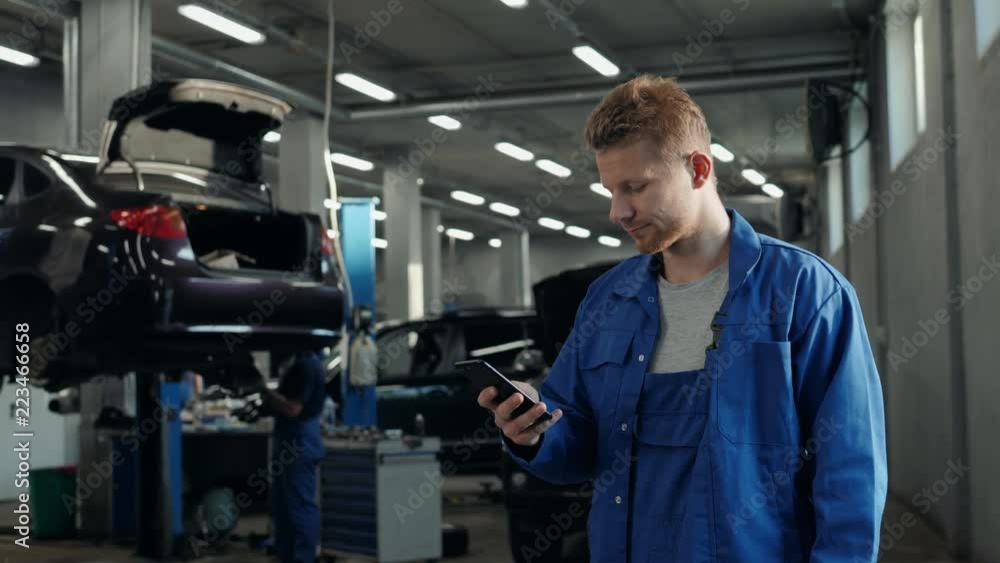 Handsome young man talking on the mobile phone and smiling while standing in workshop with car in the background mechanic Handsome young man talking on the mobile phone and smiling while standing in workshop with car in the background mechanic