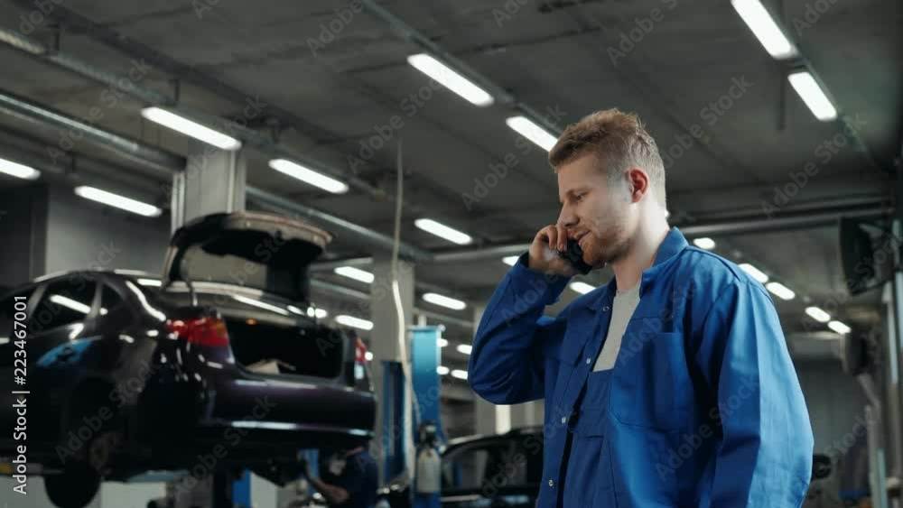 Handsome young man talking on the mobile phone and smiling while standing in workshop with car in the background mechanic Handsome young man talking on the mobile phone and smiling while standing in workshop with car in the background mechanic
