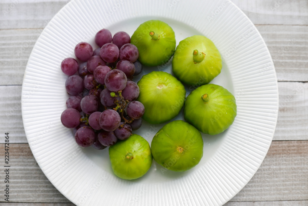 Green figs and purple grapes in a white bowl centered on a old wood table