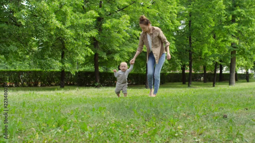 Sideways tracking shot of cheerful young mother holding hand of upset baby boy in overalls and helping him walk on green grass in park on summer day Sideways tracking shot of cheerful young mother holding hand of upset baby boy in overalls and helping him walk on green grass in park on summer day