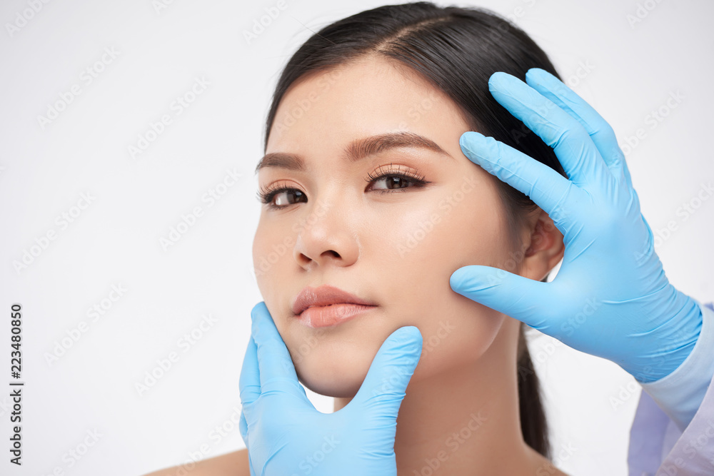 Gloved hands of cosmetologist examining flawless face of beautiful young woman Gloved hands of cosmetologist examining flawless face of beautiful young woman