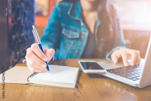 A woman's hand is writing on a notepad with a pen and a phone with a laptop.