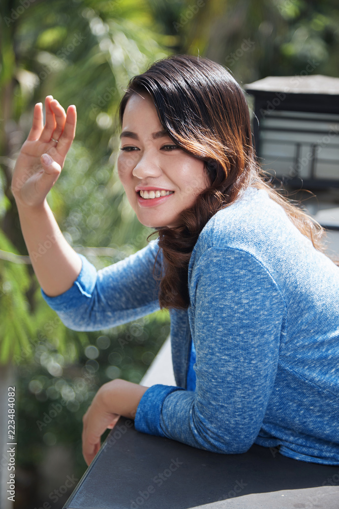 Attractive smiling Vietnamese woman standing on balcony and waving somebody Attractive smiling Vietnamese woman standing on balcony and waving somebody