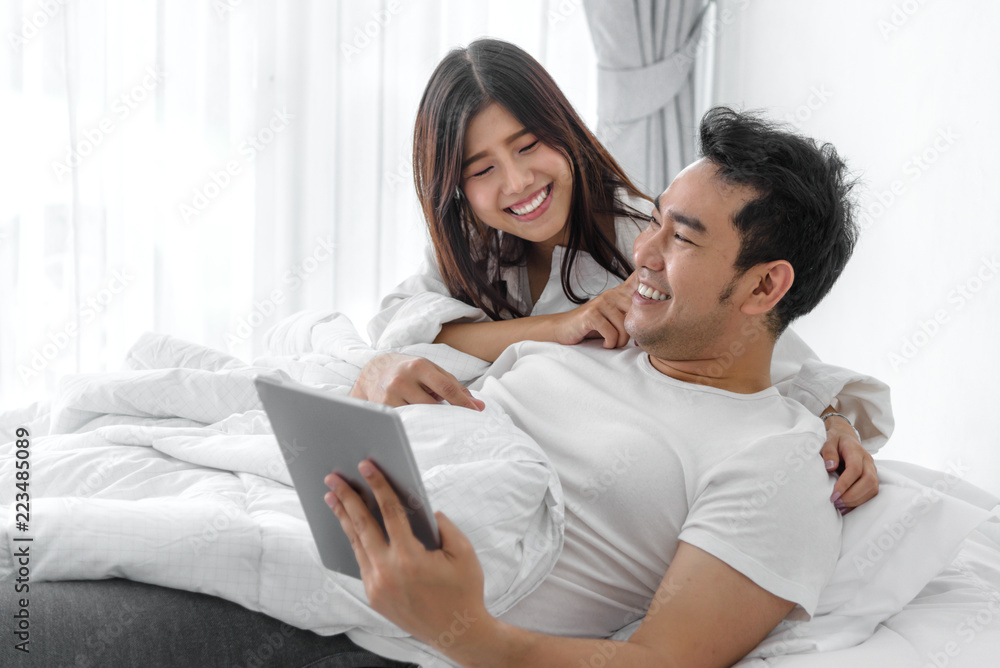 Happy Asian couple using laptop and tablet together on bed at home, lifestyle concept.