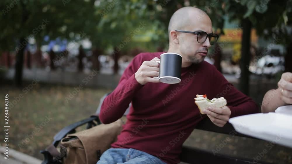 Two men having lunch on a Park bench with a sandwich and drink from cups Two men having lunch on a Park bench with a sandwich and drink from cups