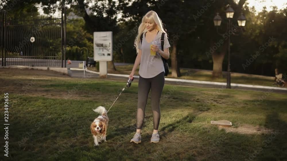 Front view of beautiful smilling woman with long blonde hair walking with her dog in the park .She is wearing casual clothes and holding drink in hand Front view of beautiful smilling woman with long blonde hair walking with her dog in the park .She is wearing casual clothes and holding drink in hand
