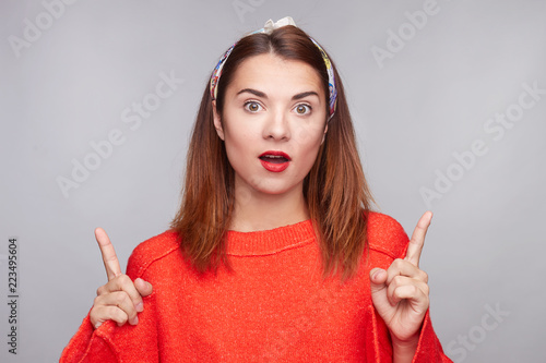 Bild auf Leinwand Studio portrait of beautiful young hipster female, stares at camera with bugged eyes, keeps jaw dropped, points with both index fingers upwards, expresses unexpectedness and shows surprise on gray