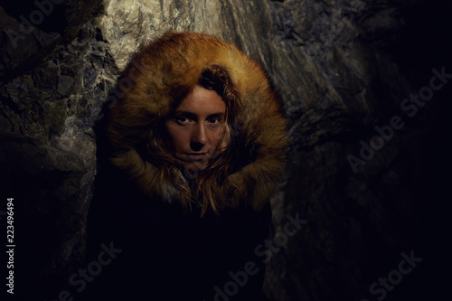 Authentic close up portrait of Caucasian female traveler, dressed in dark coat with foxy fur hood, hiding in cave from thunder, spending vacations in mountains hiking. Tourism and exploring concept. © Yulia