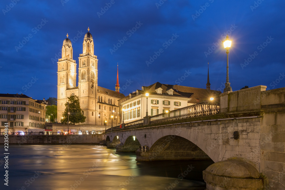 Beautiful cityscape of Zurich, Switzerland, with the Grossmünster Protestant church and the Helmhaus museum, seen from the shore of the river Limmat in the blue hour in summer
