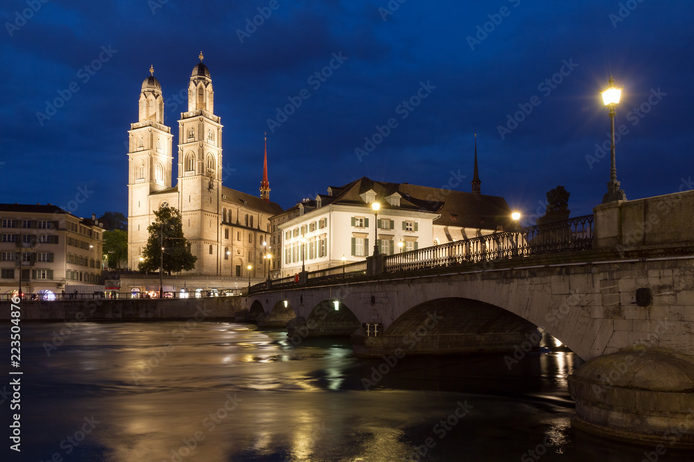 Beautiful cityscape of Zurich, Switzerland, with the Grossmünster Protestant church and the Helmhaus museum, seen from the shore of the river Limmat in the blue hour in summer
