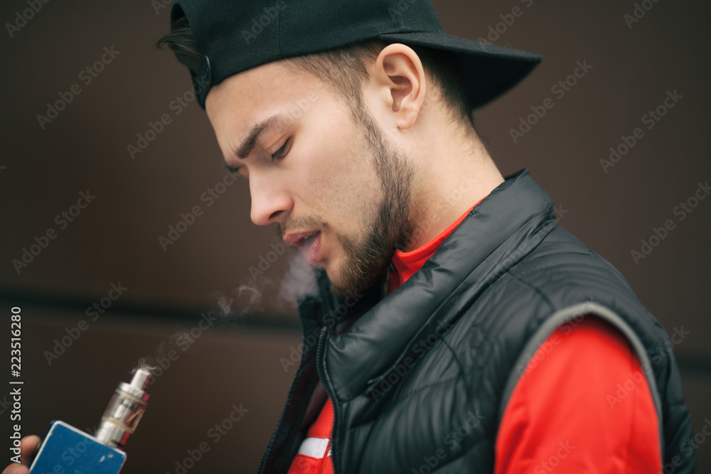 Vape teenager. Young handsome bearded white man in black cap smoking an electronic cigarette opposite the modern brown background in autumn. Vape teenager. Young handsome bearded white man in black cap smoking an electronic cigarette opposite the modern brown background in autumn.