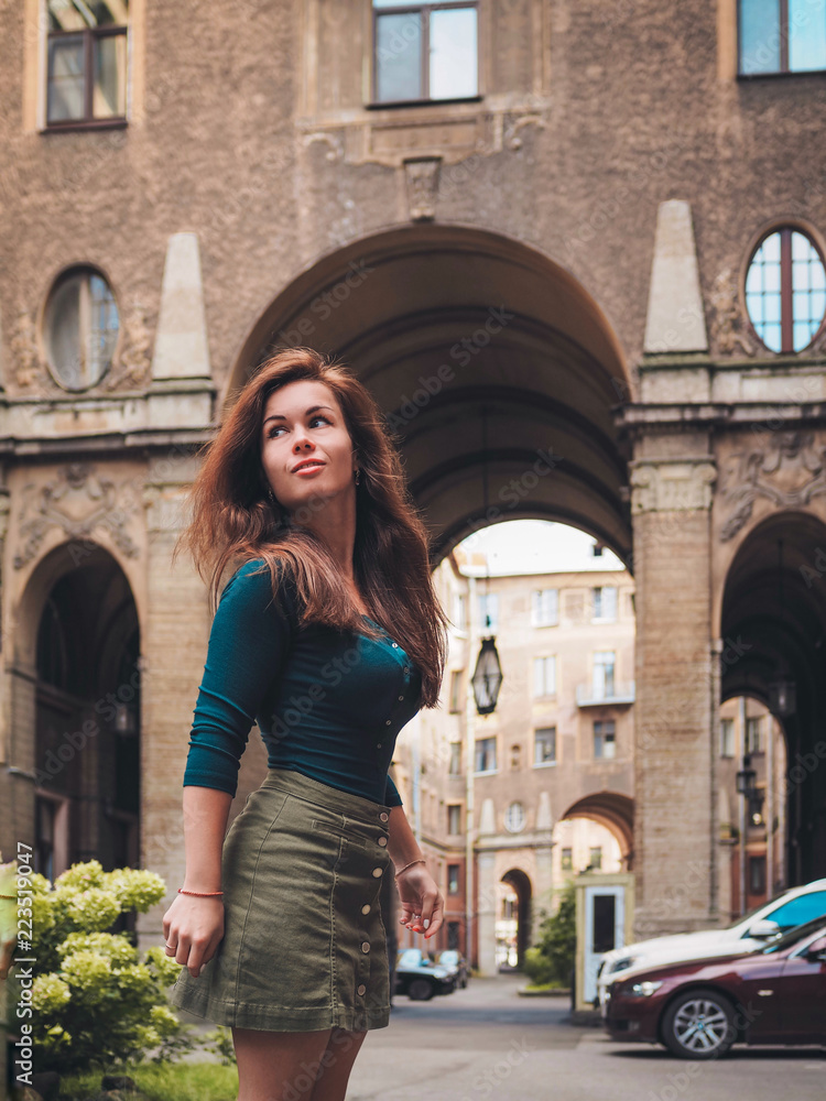Fototapeta premium Brunette girl in a skirt and with long hair, standing, posing in the courtyard of a historic building in St. Petersburg, the entrance of the house, the house is made of numerous huge arches
