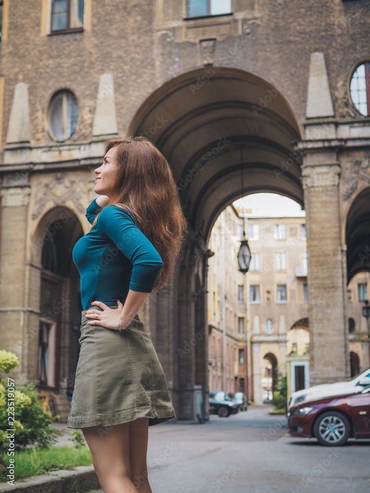 Brunette girl in a skirt and with long hair, standing, posing in the courtyard of a historic building in St. Petersburg, the entrance of the house, the house is made of numerous huge arches