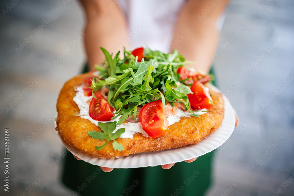 Fototapeta premium A young woman holding a traditional Hungarian Langos with cheese, rucola and tomatoes , Budapest, Hungury