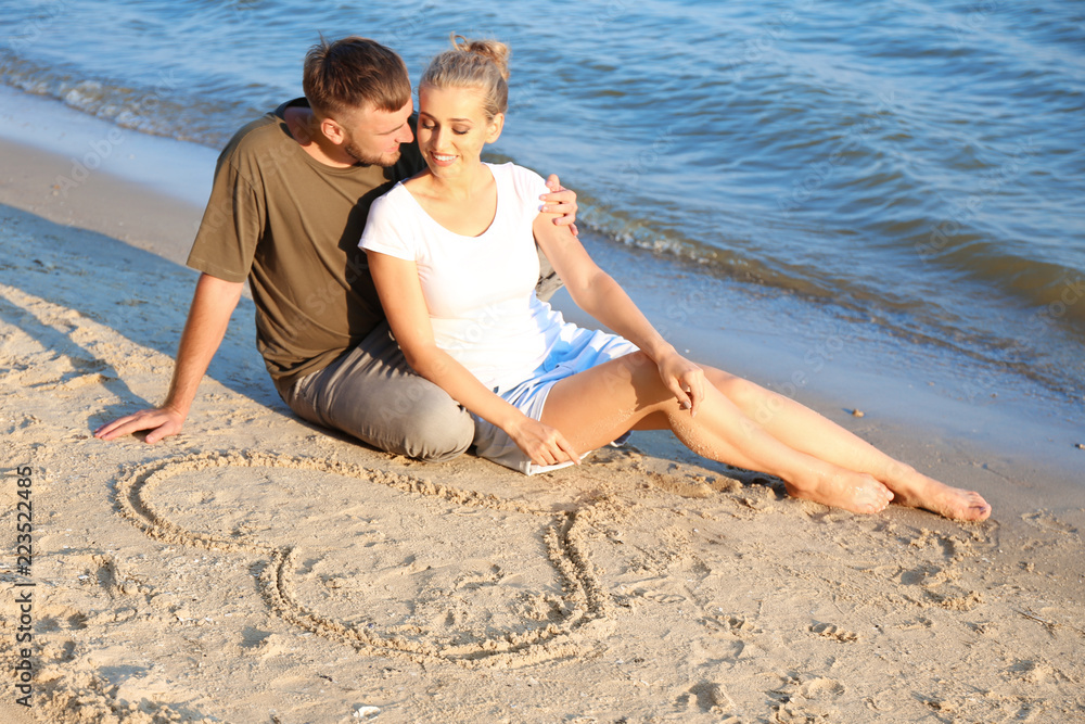 Cute young couple sitting near heart drawn on beach sand Cute young couple sitting near heart drawn on beach sand