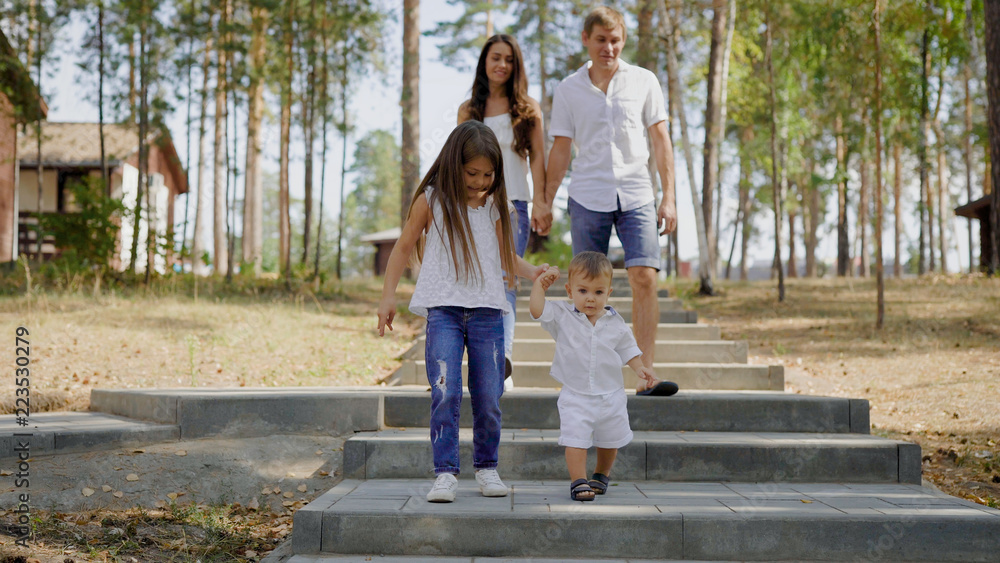 Obraz premium little girl is holding hands of her baby brother, walking together on stone stair in a park in daytime, parents are behind
