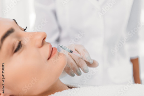 cropped shot of cosmetologist in latex glove with syringe making beauty injection to beautiful woman in spa salon