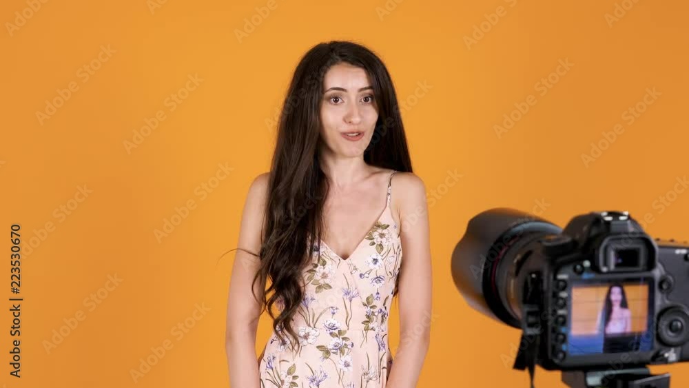 Gorgeous woman in summer dress vlogging in front of DSLR camera. There is an orange yellow background and she is an a studio Gorgeous woman in summer dress vlogging in front of DSLR camera. There is an orange yellow background and she is an a studio