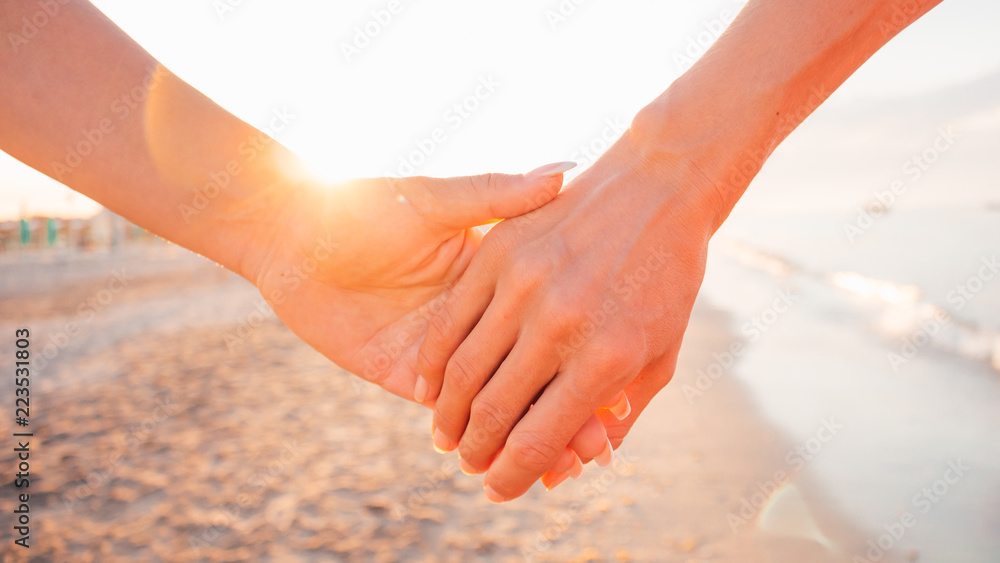 Close up of two female holding hands against sunset - two girls walking on the beach hand by hand - same-sex lesbian couple and female friendship concepts