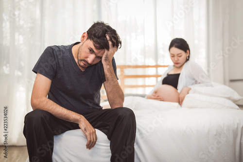 Worried stress man sitting on bed with hand on forehead in bedroom in serious mood emotion with pregnant wife woman background. Major Depressive Disorder called MDD concept. Physical healthcare