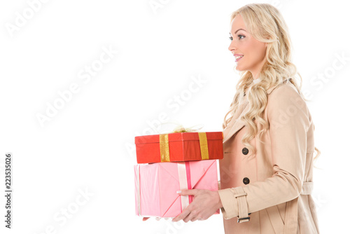 smiling blonde woman in beige coat holding gifts, isolated on white