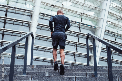 Move your body! Full length rear view of young man in sportswear jogging on staircase against industrial city view