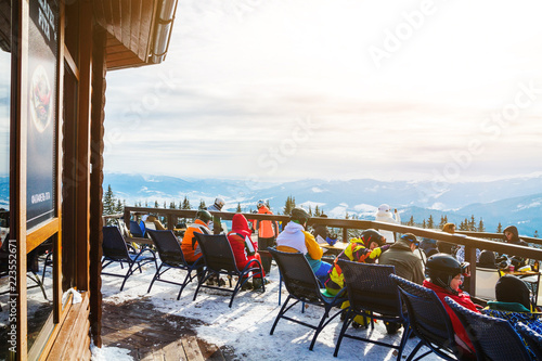 skiers sit on chairs near a restaurant on top of a mountain. People in ski suits rest and drink