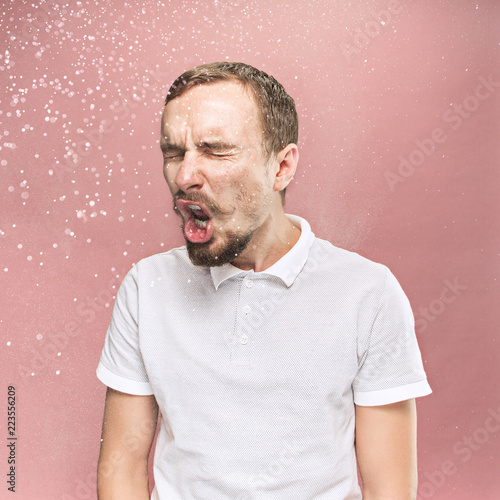 Young funny handsome man with beard and mustache sneezing with spray and small drops, studio portrait on pink background. Comic, caricature, humor. illness, infection, ache. Health concept photo