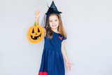 cheerful happy little girl with red horns and pumpkin stands on a gray background. selective focus.	