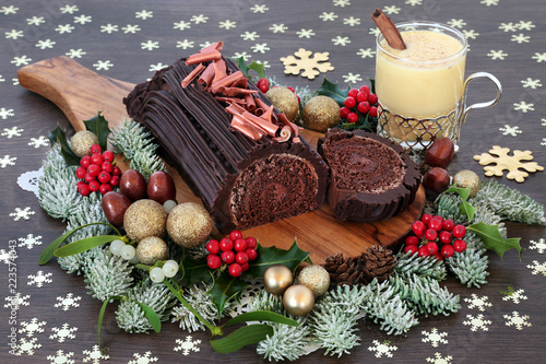Chocolate yule cake with eggnog, traditional winter flora, gold baubles and snowflake decorations on rustic oak wood background. Festive sweet food for Christmas.