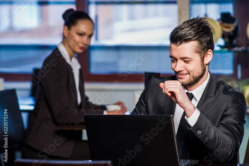 Handsome young man working on laptop and smiling while enjoying coffee in cafe with yang woman in background.