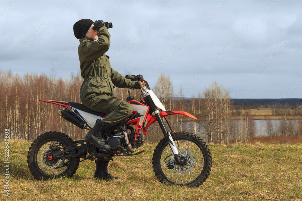 Caucasian girl is on a motorcycle with binoculars. The cross bike is in  outdoors. The biker