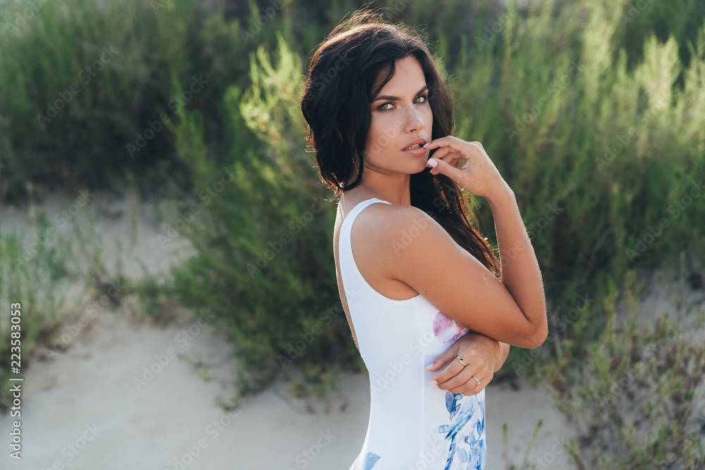 Young stylish brunette has a European appearance, wears a trendy white swimsuit, poses sideways against the background of sand on an abandoned empty beach, touches her lips with her finger. Young stylish brunette has a European appearance, wears a trendy white swimsuit, poses sideways against the background of sand on an abandoned empty beach, touches her lips with her finger.