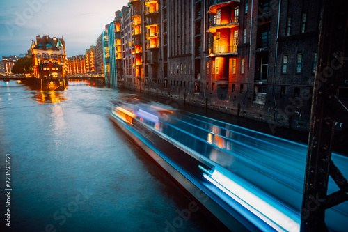 A colorfully illuminated boat cruising on the Wandrahmsfleet at night. The Warehouse District in Hamburg, Germany. The district located within the HafenCity quarter. Long exposure photo