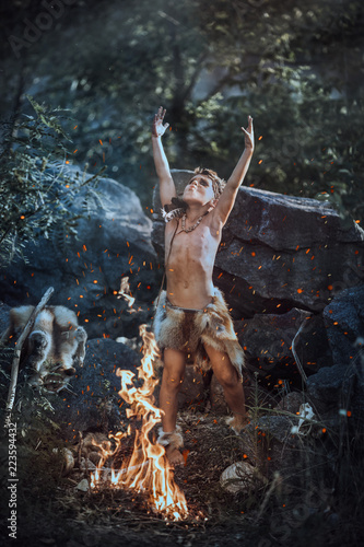 Shaman boy at the fire. Scary young primitive boy outdoors near bonfire. Witch craft concept. Angry caveman, manly boy with horns near bonfire. Prehistoric tribal man outdoors on nature making