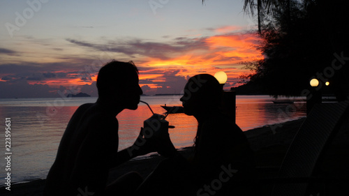 A loving couple sits on the seashore in a view of the sunset, they drink one coconut for two through a straw. HD