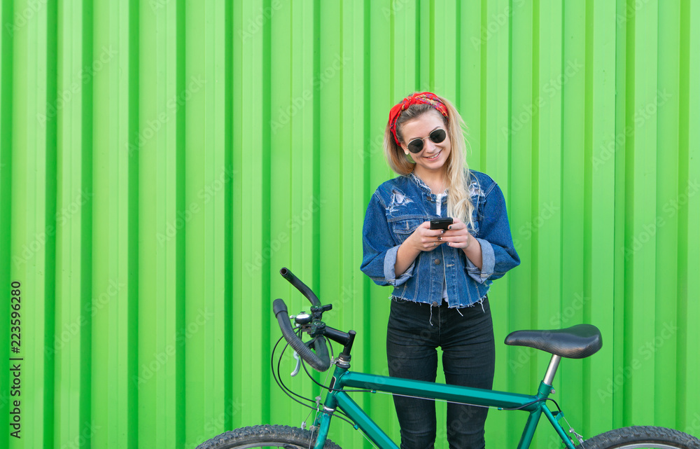 Stylish smiling girl stands in a denim jacket with a bicycle and uses a smartphone on the background of a green wall. Sports and Technology. Copyspace